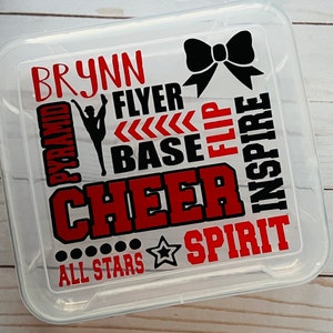 May include: Clear plastic storage container with a red and black design. The design includes the words "BRYNN", "PYRAMID", "FLYER", "BASE", "FLIP", "CHEER", "INSPIRE", "ALL STARS", and "SPIRIT". There is also a black bow and a silhouette of a cheerleader.