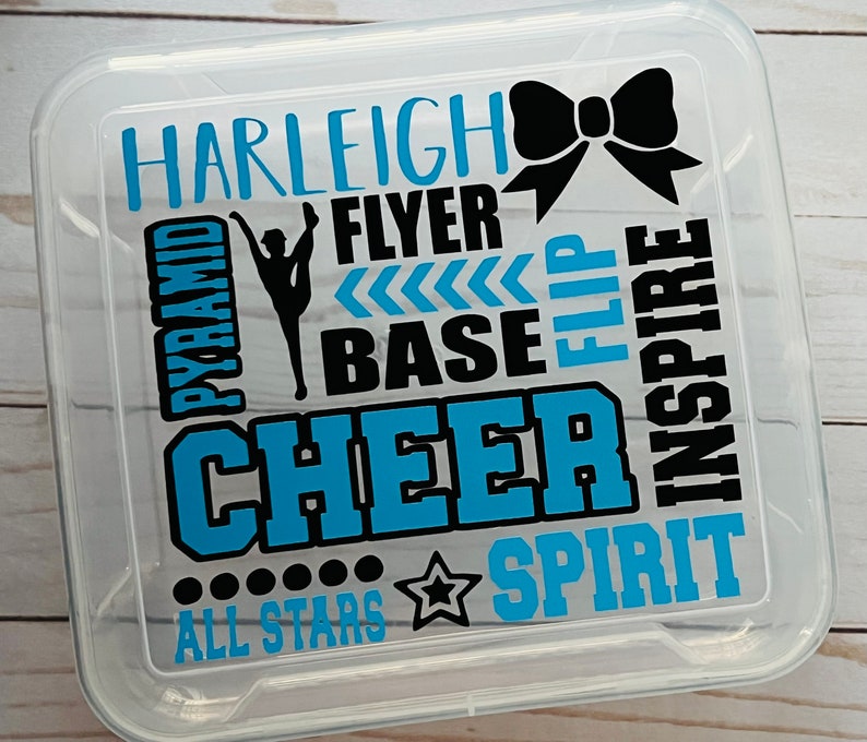 May include: Clear plastic storage box with a blue and black design. The design includes the words "Harleigh Cheer", "Pyramid", "Flyer", "Base", "Flip", "Inspire", "Spirit", and "All Stars".