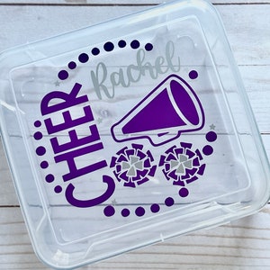 Puede incluir: Un recipiente de almacenamiento de pl&aacute;stico transparente con un dise&ntilde;o morado y plateado. El dise&ntilde;o incluye la palabra "CHEER" y el nombre "Rachel", junto con un meg&aacute;fono morado y dos pompones plateados.