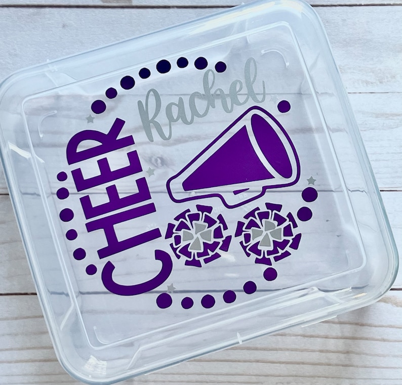 Puede incluir: Un recipiente de almacenamiento de pl&aacute;stico transparente con un dise&ntilde;o morado y plateado. El dise&ntilde;o incluye la palabra "CHEER" y el nombre "Rachel", junto con un meg&aacute;fono morado y dos pompones plateados.