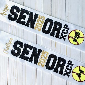 Op de afbeelding: Twee witte satijnen linten met zwarte glittertekst die "Senior 2025" en "XC/P" zegt met een gele softbal met zwarte stiksels en het nummer "#25" erop. De linten hebben de namen "Alycia" en "Chelsea" erop geschreven.