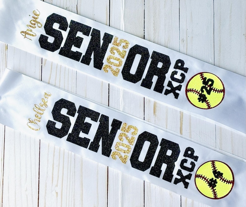 Op de afbeelding: Twee witte satijnen linten met zwarte glittertekst die "Senior 2025" en "XC/P" zegt met een gele softbal met zwarte stiksels en het nummer "#25" erop. De linten hebben de namen "Alycia" en "Chelsea" erop geschreven.