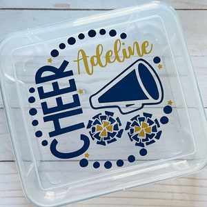 Puede incluir: Un contenedor de almacenamiento de pl&aacute;stico transparente con un dise&ntilde;o azul marino y dorado. El dise&ntilde;o presenta las palabras "CHEER" y "Adeline" junto con un meg&aacute;fono y pompones de animadora.