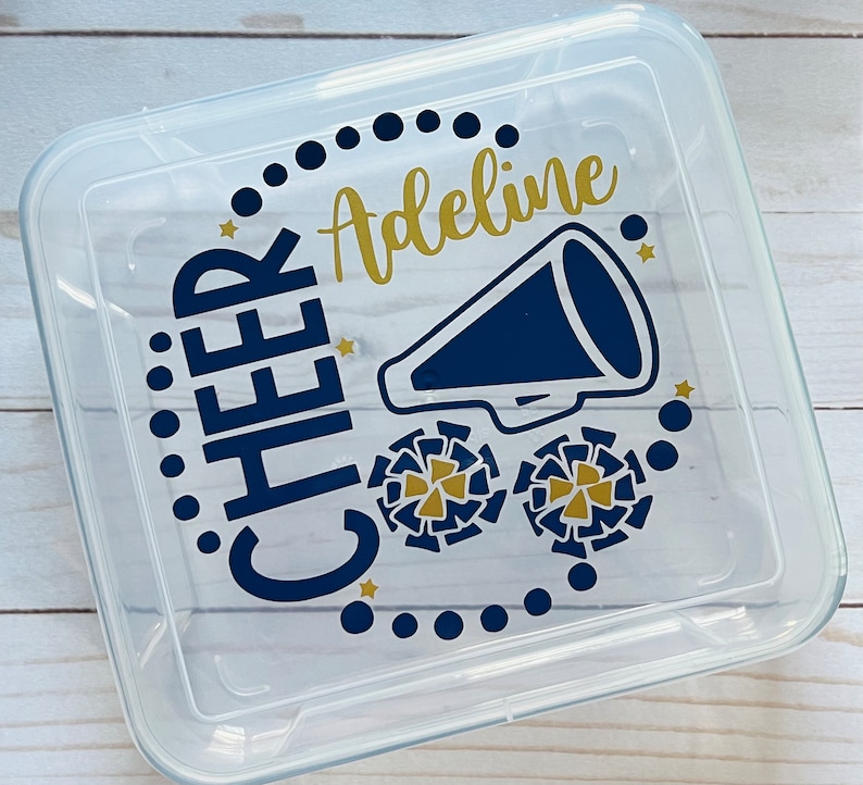 Puede incluir: Un contenedor de almacenamiento de pl&aacute;stico transparente con un dise&ntilde;o azul marino y dorado. El dise&ntilde;o presenta las palabras "CHEER" y "Adeline" junto con un meg&aacute;fono y pompones de animadora.
