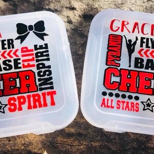 May include: Two clear plastic storage containers with black and red cheerleading themed designs. The containers have the names "Kayla" and "Grace" on them. The designs include the words "flyer", "base", "pyramid", "flip", "cheer", "inspire", "spirit", and "all stars".