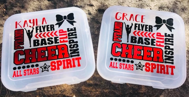May include: Two clear plastic storage containers with black and red cheerleading themed designs. The containers have the names "Kayla" and "Grace" on them. The designs include the words "flyer", "base", "pyramid", "flip", "cheer", "inspire", "spirit", and "all stars".
