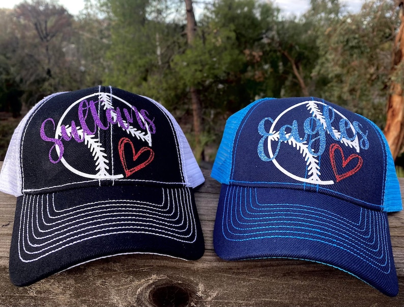 May include: Two black baseball caps with white mesh back panels. The front of each cap features a white peace sign with a baseball inside. The word "Sultans" is embroidered in purple on one cap and "Eagles" is embroidered in blue on the other cap. Both caps have a red heart embroidered on the peace sign.