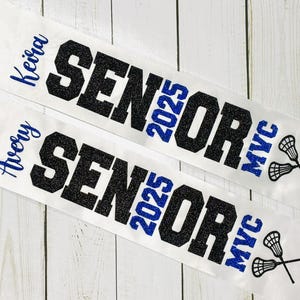 Op de afbeelding: Twee witte sjerpen met zwarte glittertekst die "Senior 2025" en "MVC" zegt. De sjerpen hebben blauwe glittertekst die "Avery" en "Keira" zegt.
