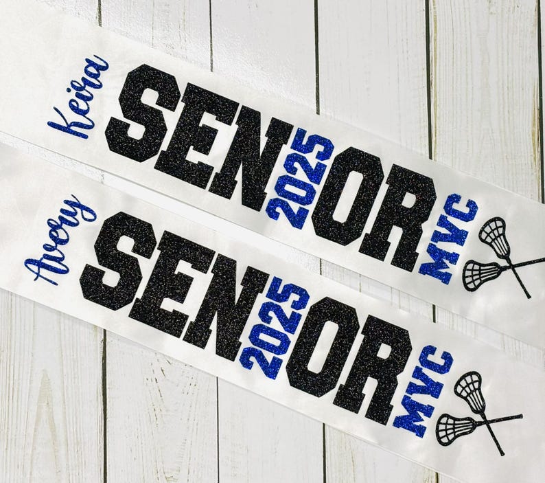Op de afbeelding: Twee witte sjerpen met zwarte glittertekst die "Senior 2025" en "MVC" zegt. De sjerpen hebben blauwe glittertekst die "Avery" en "Keira" zegt.