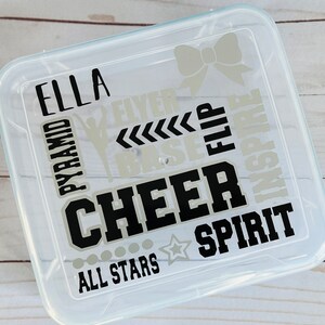 May include: Clear plastic storage container with a black and silver design. The design includes the words "Ella", "Cheer", "Spirit", "All Stars", "Pyramid", "Flyer", "Base", "Flip", and "Inspire".