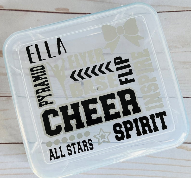 May include: Clear plastic storage container with a black and silver design. The design includes the words "Ella", "Cheer", "Spirit", "All Stars", "Pyramid", "Flyer", "Base", "Flip", and "Inspire".
