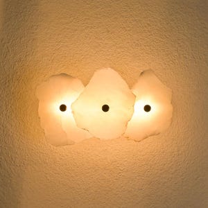 Può includere: Un'applique da parete con tre paralumi bianchi incandescenti di forma irregolare. Ogni paralume ha un centro circolare scuro, che emette una luce calda. L'apparecchio è montato su una parete strutturata bianco sporco.