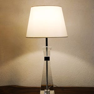 Può includere: Una lampada da tavolo in cristallo trasparente con paralume bianco. La lampada ha una base geometrica e un accento nero. Il paralume ha una forma conica classica. La lampada è su una superficie di legno.