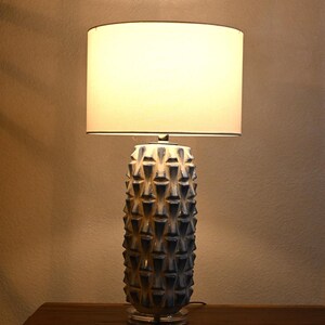 Puede incluir: Una lámpara de mesa decorativa con una pantalla blanca y una base de cerámica azul y blanca texturizada. La lámpara es cilíndrica con un patrón geométrico repetitivo. La lámpara está sobre una superficie de madera.