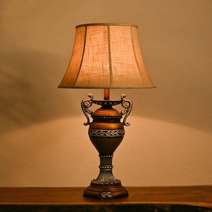 May include: A decorative table lamp with a bronze-colored base and a beige lampshade. The base features ornate detailing and a classic urn shape. The lampshade is a light tan color, and the lamp is sitting on a wooden surface.