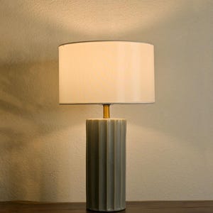 Può includere: Una lampada da tavolo in ceramica grigia con paralume bianco. La lampada ha una base cilindrica con scanalature verticali. Il paralume è rettangolare e ha un bordo grigio scuro. La lampada è su una superficie di legno marrone scuro.