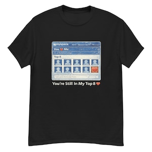 May include: Black t-shirt featuring a nostalgic graphic of a MySpace profile, with the text "You ❤️ Me" and "You're Still In My Top 8." The design includes a heart icon and profile pictures, evoking a retro internet theme.