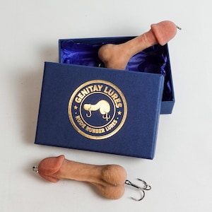 May include: Two novelty fishing lures in the shape of a penis, one inside a navy blue box with gold lettering and a logo that reads "GENITAY LURES RUDE RUBBER LURES". The lures are flesh-toned with a silver hook.