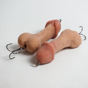 May include: Two novelty fishing lures shaped like penises. The lures are flesh-toned with a darker pink head and a black fishing hook attached. One lure has a small metal ring attached.