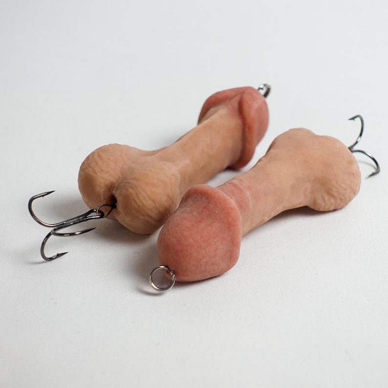 May include: Two novelty fishing lures shaped like penises. The lures are flesh-toned with a darker pink head and a black fishing hook attached. One lure has a small metal ring attached.