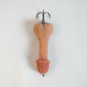 May include: A novelty fishing lure in the shape of a penis, with a dark metal treble hook attached at the top and a small hook at the bottom. The lure is a flesh-toned color with a darker reddish-brown tip.