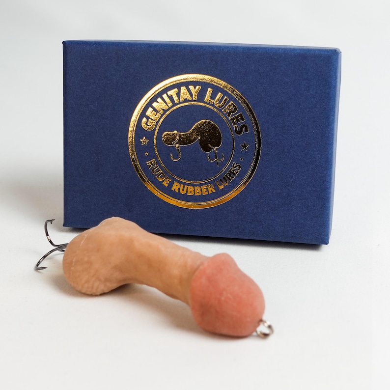 May include: A novelty fishing lure shaped like a penis, in a flesh-tone color, with a pink head and a silver hook. The lure is in front of a navy blue box with gold lettering that reads "GENITAY LURES" and "RUDE RUBBER LURES".