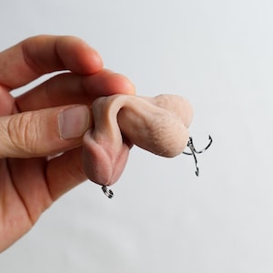 May include: A flesh-toned sculpture of male genitalia with a metal hook attached. The sculpture is held in a hand against a white background. The hook is a fishing lure.