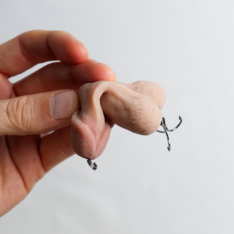 May include: A flesh-toned sculpture of male genitalia with a metal hook attached. The sculpture is held in a hand against a white background. The hook is a fishing lure.