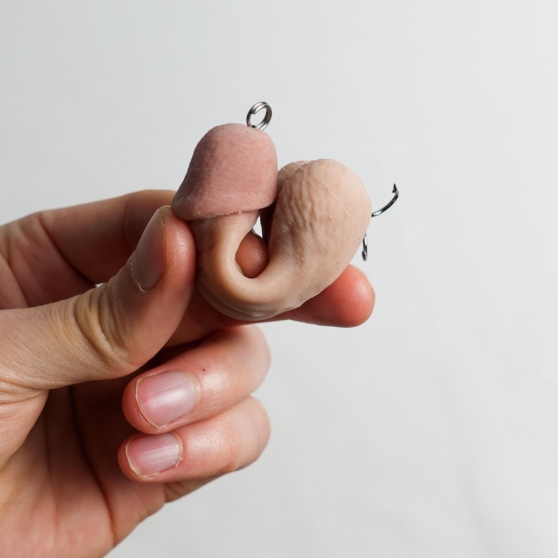 May include: A small, flesh-toned sculpture of a stylized anatomical form, featuring a rounded head and a curved body. The sculpture has a textured surface and a small metal loop for hanging. The piece is held in a hand against a plain white background.