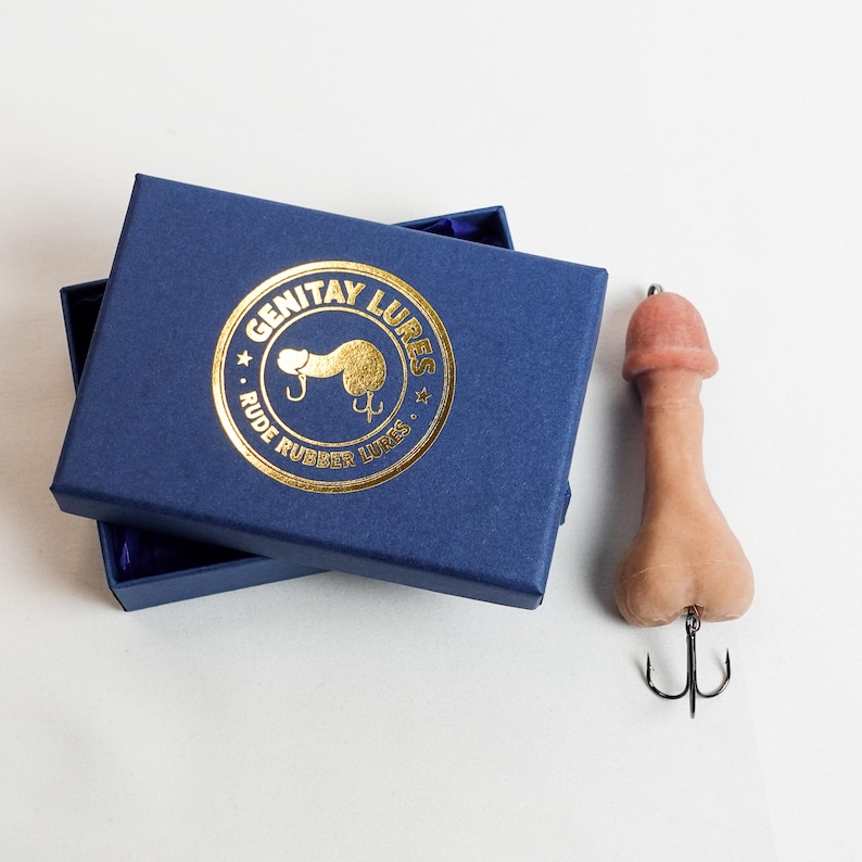 May include: A novelty fishing lure shaped like a penis, in shades of pink and tan, with a black treble hook. It is next to a navy blue box with gold lettering that reads "Genitay Lures" and "Rude Rubber Lures".
