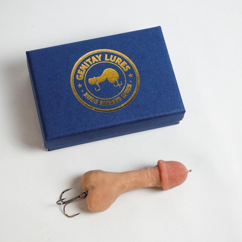 May include: A novelty fishing lure shaped like a penis, with a hook attached. The lure is flesh-toned with a pink head. It is next to a blue box with gold text that reads "GENITAY LURES" and "RUDE RUBBER LURES".