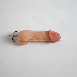 May include: A novelty fishing lure shaped like a penis. The lure is flesh-toned with a pink head and a metal hook attached. The design is humorous and unconventional.