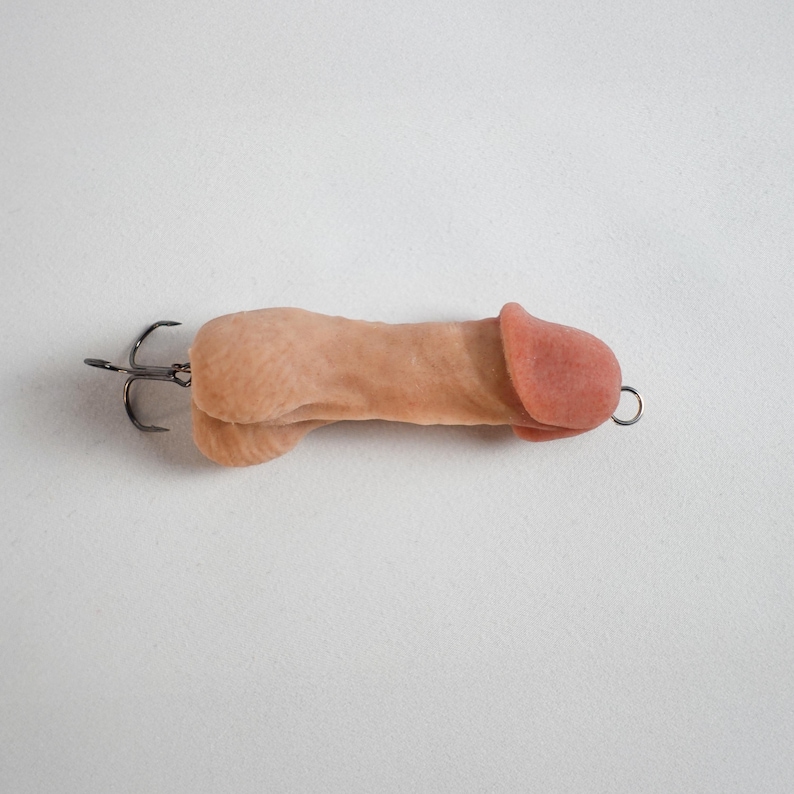 May include: A novelty fishing lure shaped like a penis. The lure is flesh-toned with a pink head and a metal hook attached. The design is humorous and unconventional.