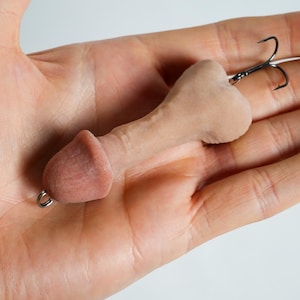 May include: A novelty fishing lure shaped like a phallus, in a flesh-toned color. It features a small metal ring at the base and a treble hook at the top. The lure is designed to attract fish.