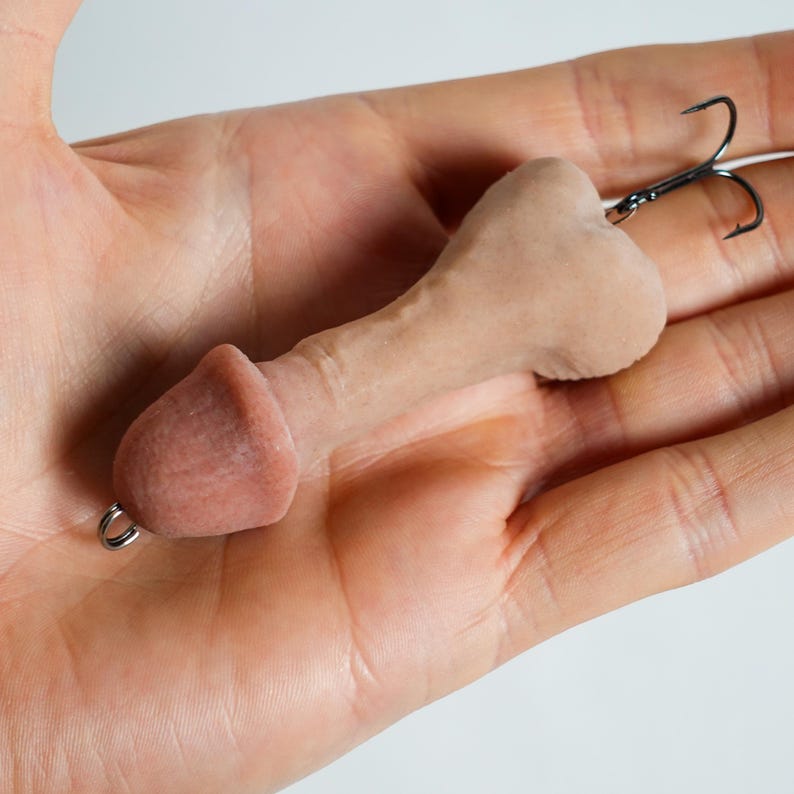 May include: A novelty fishing lure shaped like a phallus, in a flesh-toned color. It features a small metal ring at the base and a treble hook at the top. The lure is designed to attract fish.