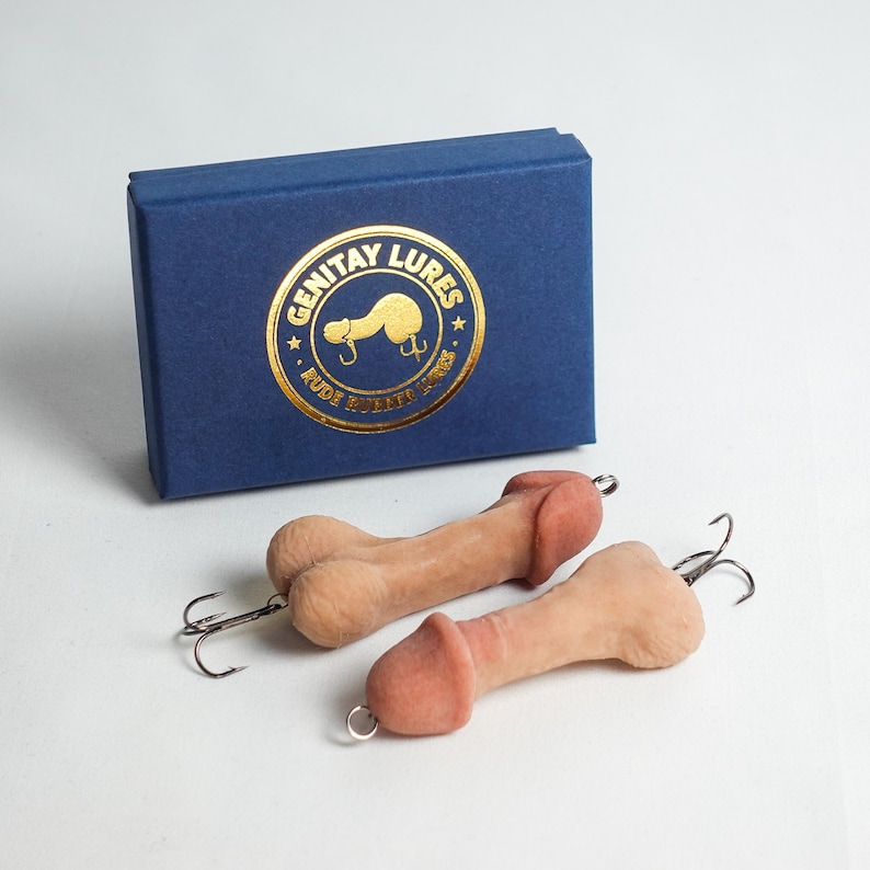 May include: Two novelty fishing lures in the shape of a penis, with a light brown body and a pink head. The lures have a silver hook and are next to a blue box with gold text that reads "Genitay Lures".
