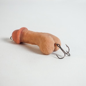May include: A novelty fishing lure shaped like a penis. The lure is a flesh-toned color with a darker head and features two black treble hooks attached to the base. The background is plain white.