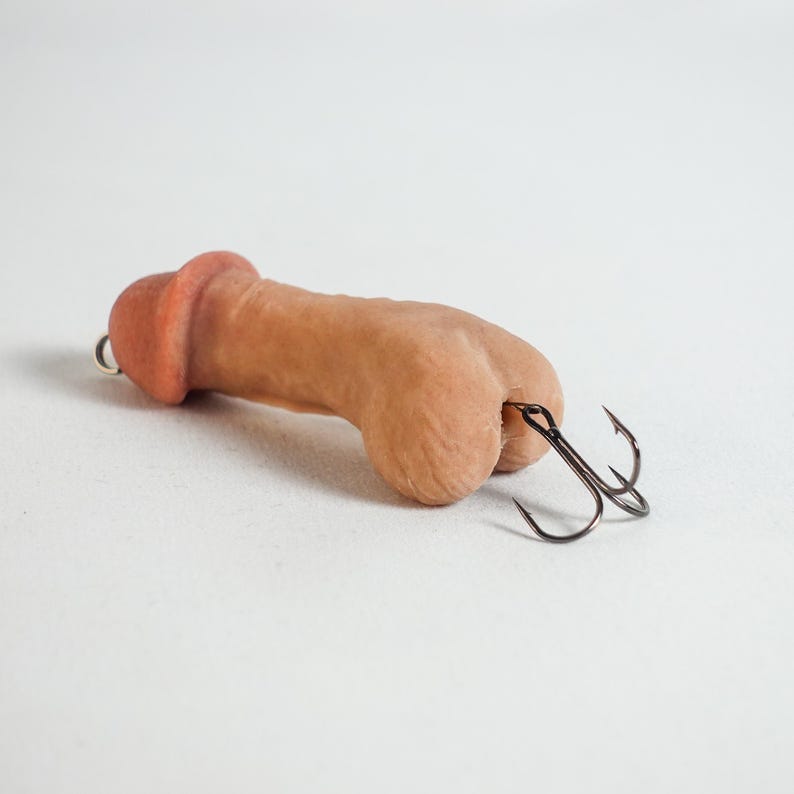 May include: A novelty fishing lure shaped like a penis. The lure is a flesh-toned color with a darker head and features two black treble hooks attached to the base. The background is plain white.