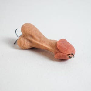 May include: A novelty item shaped like a penis, in a flesh-tone color. The item has a small metal ring attached to the tip and a black hook. The background is plain white.