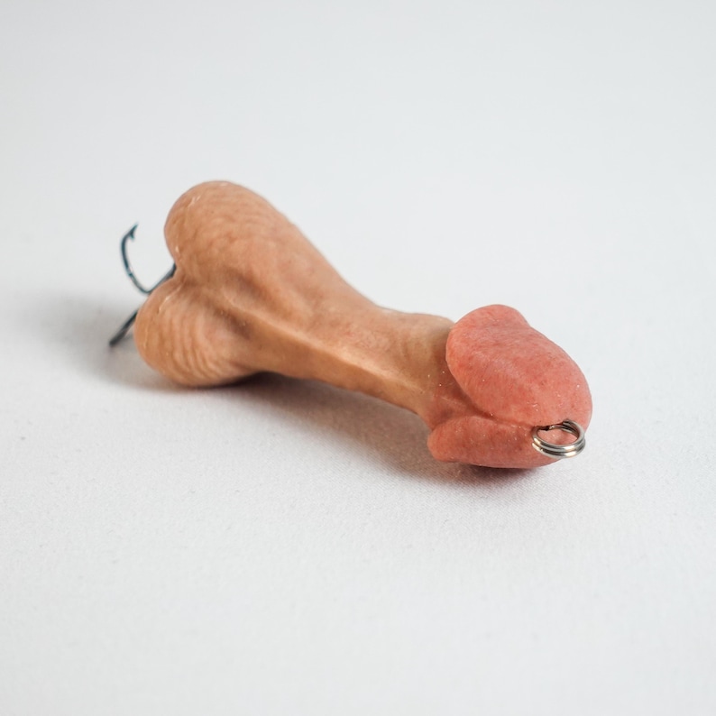 May include: A novelty item shaped like a penis, in a flesh-tone color. The item has a small metal ring attached to the tip and a black hook. The background is plain white.