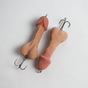 May include: Two novelty fishing lures shaped like penises. The lures are flesh-toned with darker pink tips and feature a treble hook attached to the base. The background is plain white.