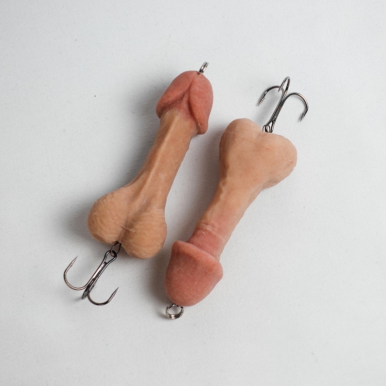 May include: Two novelty fishing lures shaped like penises. The lures are flesh-toned with darker pink tips and feature a treble hook attached to the base. The background is plain white.