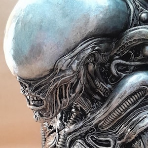 Puede incluir: Escultura detallada de una criatura alienígena, con una cabeza plateada lisa y un cuerpo texturizado intrincado. El diseño muestra una estética biomecánica con elementos acanalados y tubulares, creando un impacto visual llamativo.