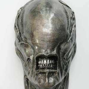 Puede incluir: Escultura plateada de una cabeza alienígena. Presenta un cráneo liso y alargado y una boca prominente con dientes. El diseño incluye detalles acanalados y una superficie texturizada, creando una estética llamativa y de otro mundo.