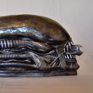 Puede incluir: Escultura gris metálica de una cabeza alienígena, con una forma ovalada alargada, detalles de crestas y una boca amenazante llena de dientes afilados. La escultura tiene una superficie texturizada y es probablemente un accesorio o artículo de colección.