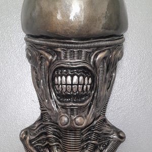 May include: A metallic sculpture of an alien head, featuring a smooth, rounded top and a detailed, textured face. The face has a wide, toothy grin and intricate mechanical details, with a silver and gray color scheme.