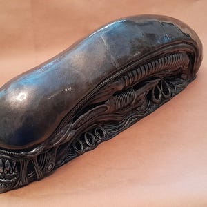 Puede incluir: Una escultura detallada de una cabeza de Alien Xenomorfo, con una cabeza larga y lisa y detalles intrincados y texturizados. La escultura es de color gris oscuro con un brillo metálico, mostrando el diseño icónico de la criatura. La boca está abierta, revelando dientes afilados.