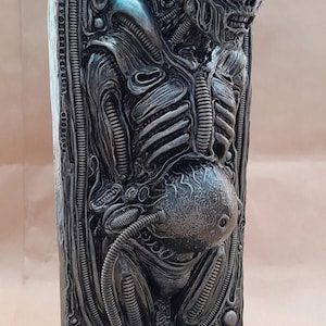 Può includere: Scultura dettagliata di una creatura aliena con testa scheletrica, torace a coste e addome bulboso. La scultura è principalmente argentata con ombre scure, che mostra intricati dettagli meccanici e organici. La base è un rettangolo arrotondato.