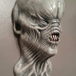 May include: A gray, sculpted wall hanging depicting a grotesque alien head. The creature has a large, bulbous head, wrinkled skin, and a menacing expression with sharp teeth. The sculpture is mounted on a gray surface.