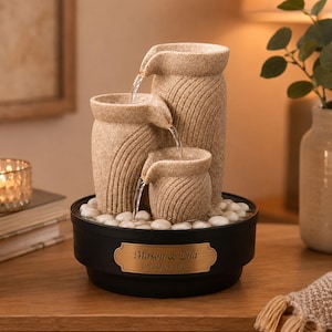 Peut inclure: Une fontaine à eau de table à trois niveaux avec une finition en pierre beige texturée et de l'eau en cascade. La base circulaire noire comporte une plaque dorée gravée du texte Mason & Lila Love Grows Here, entourée de galets blancs.